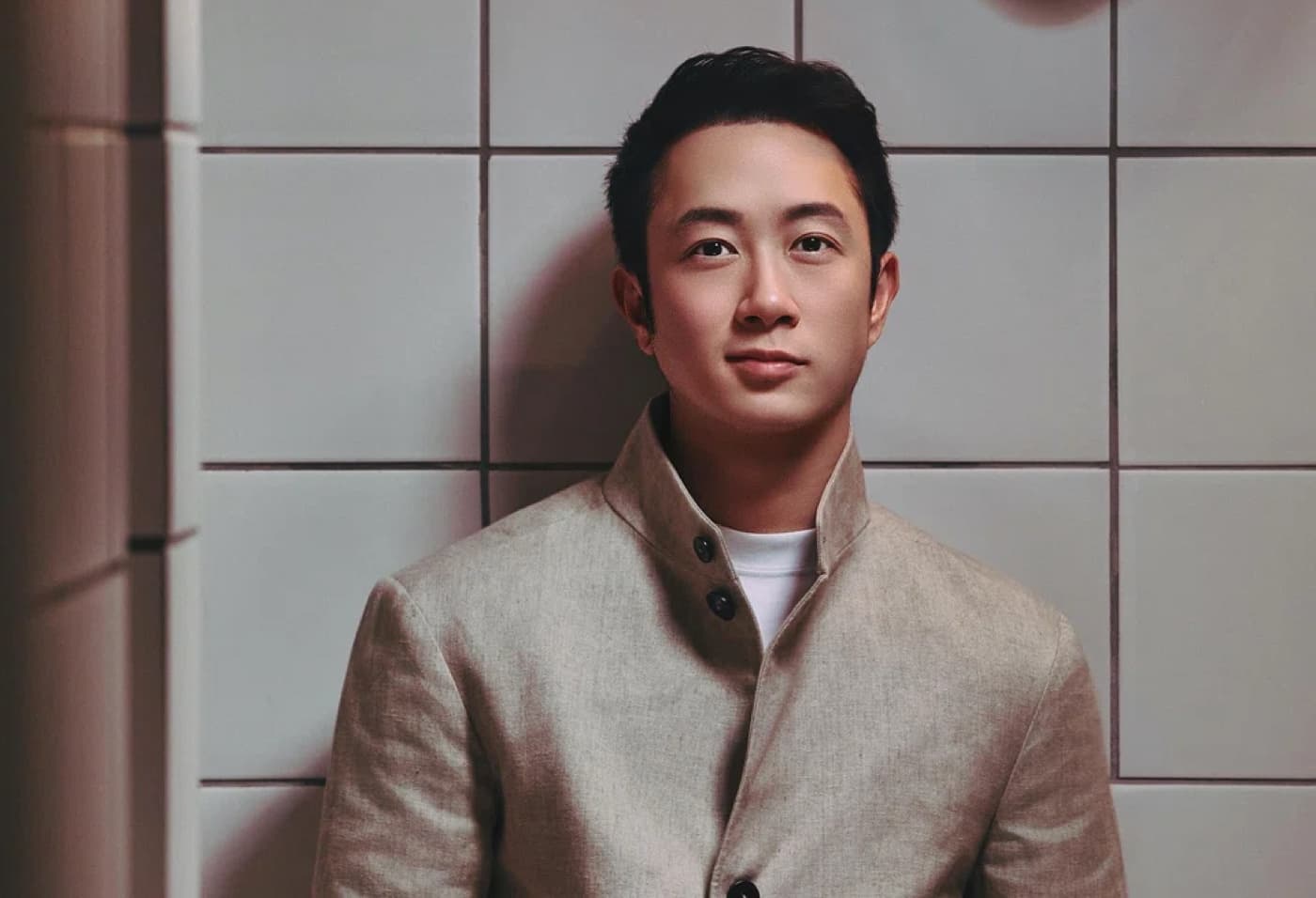 Grant Wee standing against a white tiled wall in a beige jacket