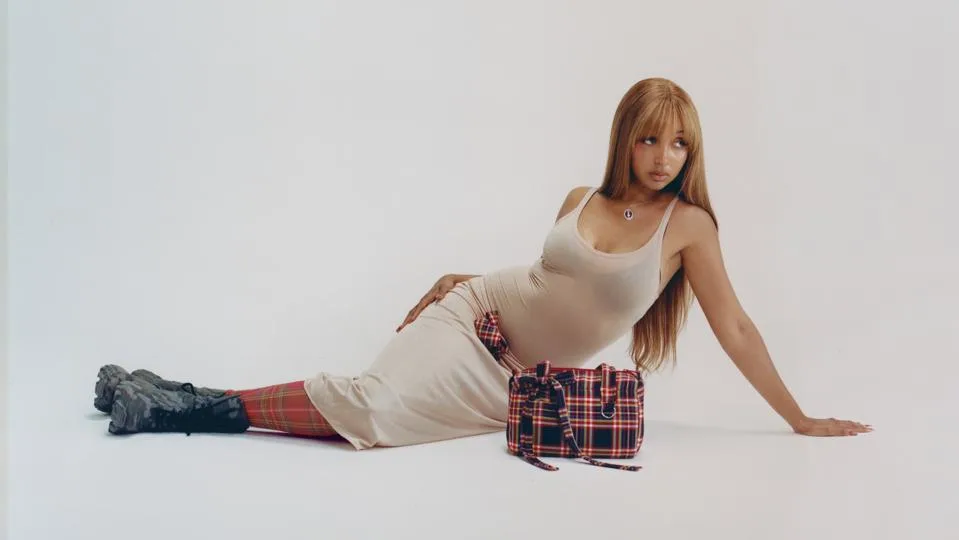 A model seated on the floor wearing a neutral tank dress, plaid socks, and combat boots beside a matching plaid handbag