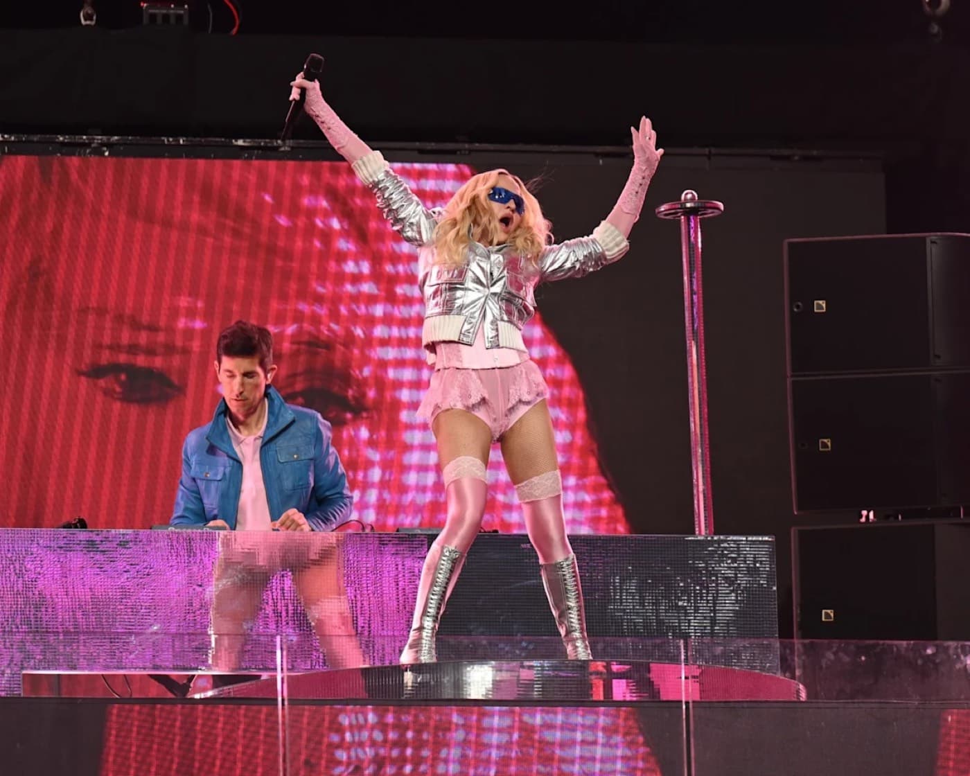 Madonna performing on stage in a pink and silver outfit with a DJ behind her
