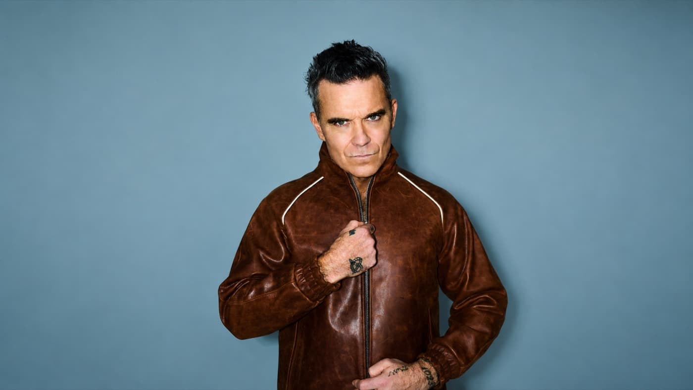 Robbie Williams in a brown leather jacket against a blue backdrop
